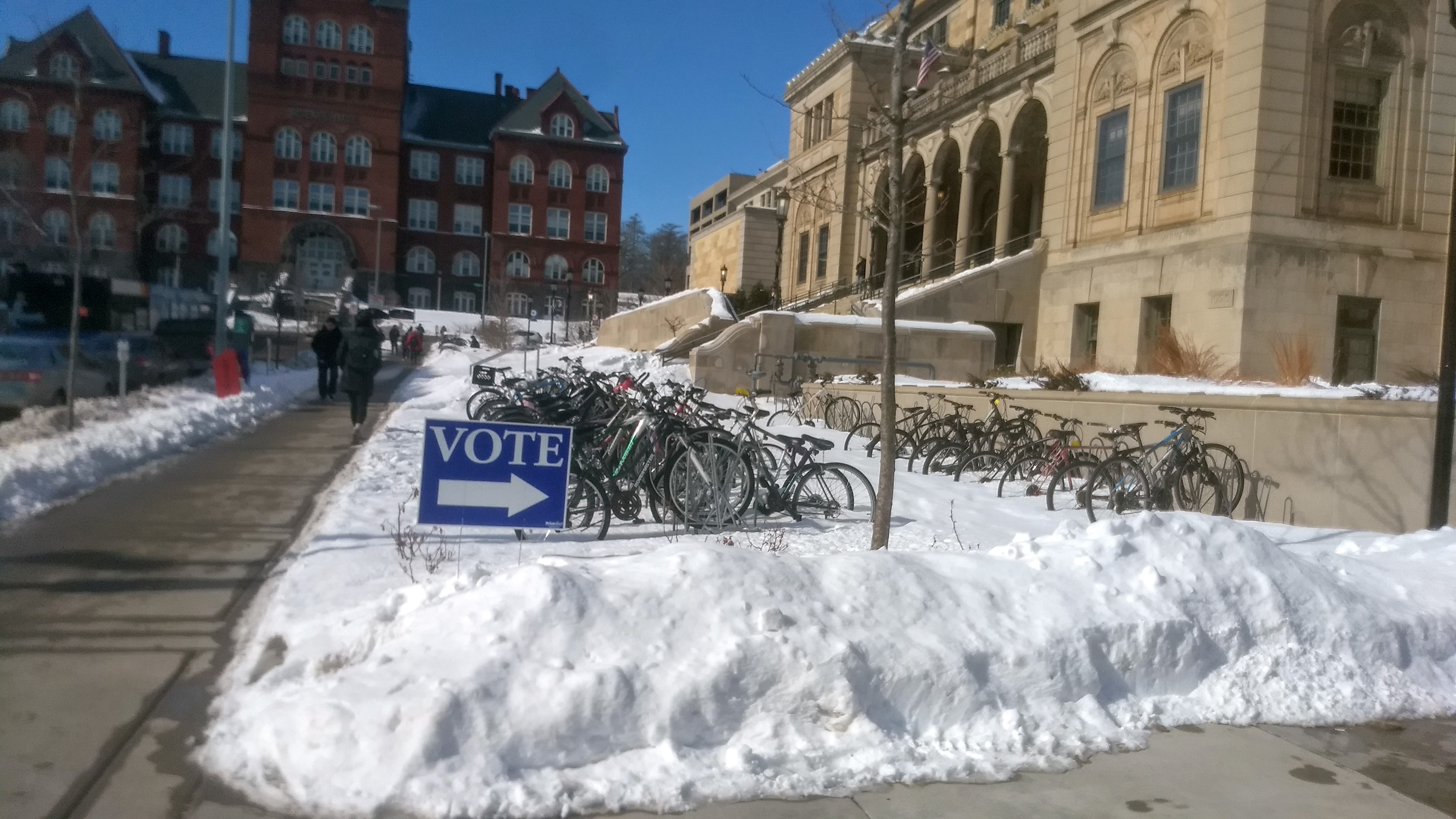 A blue yard sign saying “Vote” with an arrow pointing right in front of Memorial Union. The arrow appears to be pointing at a number of bike racks covered in snow but still well occupied with bikes.